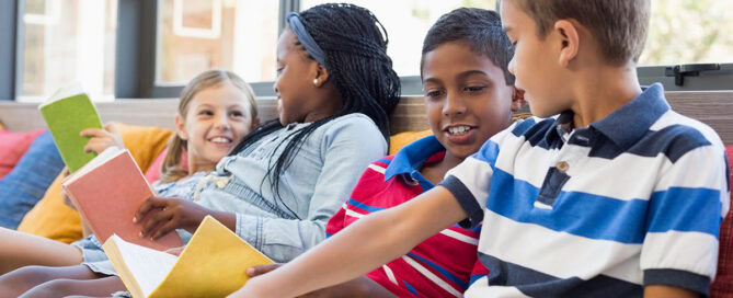 Four children sit on a couch, joyfully reading books of various colors. One girl giggles while looking at another girl, who has braids and is facing away. Two boys appear engaged in conversation, sharing a book between them. The setting is bright and inviting, with colorful cushions surrounding them.