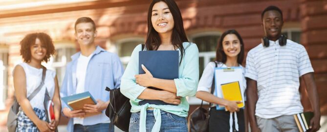 A diverse group of five students stands outside a university building, smiling at the camera. The central student, a young woman with long dark hair, holds a blue folder and wears a light green blouse. The others are dressed casually and carry books and bags.