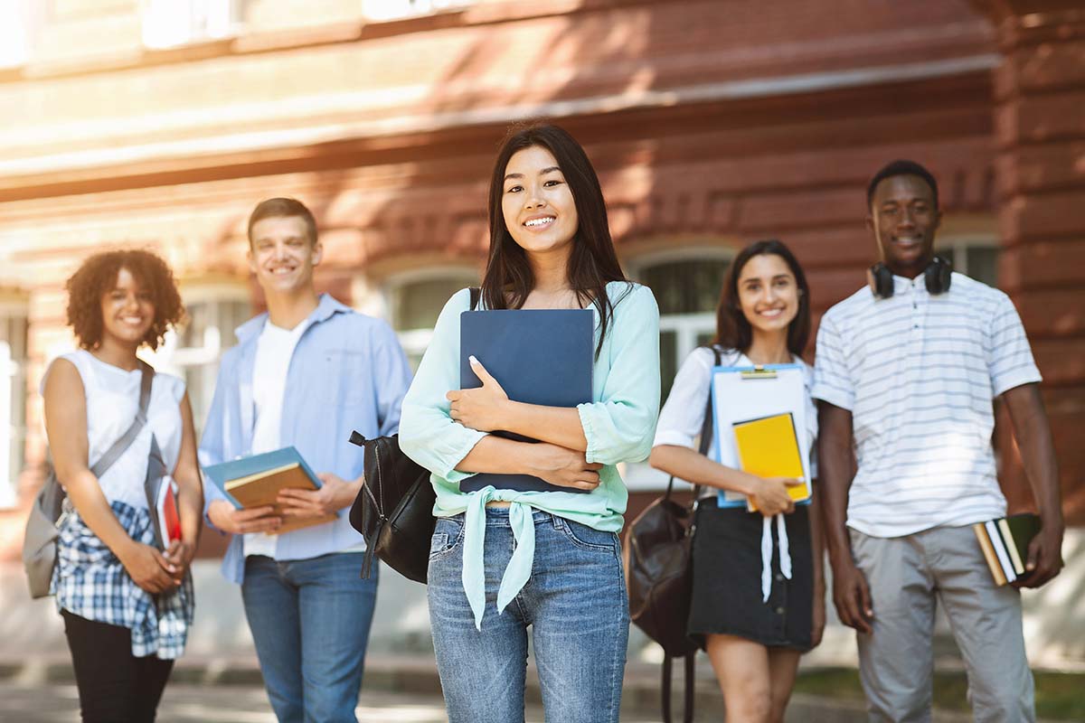 College Students on Campus A diverse group of five students stands outside a university building, smiling at the camera. The central student, a young woman with long dark hair, holds a blue folder and wears a light green blouse. The others are dressed casually and carry books and bags.