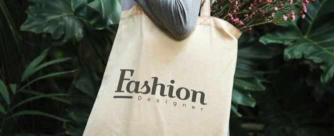 A person wearing a gray long-sleeve shirt is holding a beige tote bag with the text "Fashion Designer" printed in bold letters. The bag contains a bouquet of pink flowers, and there are lush green plants in the background.