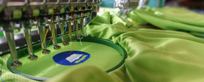 A close-up view of an embroidery machine with needle heads and colorful threads, stitching a blue logo on a green fabric.