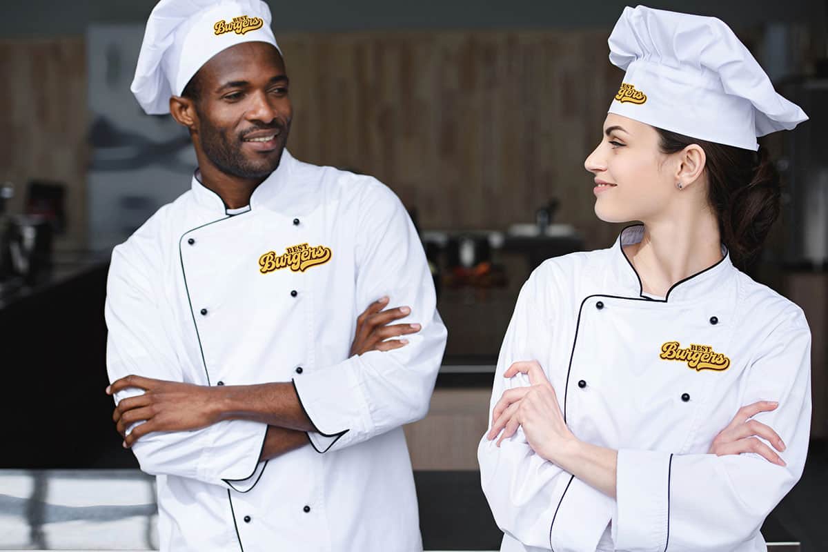 Branded Chef Uniforms Two chefs, a man and a woman, are standing in a kitchen. Both wear white chef jackets with the logo "Best Burgers" and chef hats. They are smiling at each other, with their arms crossed, conveying a friendly and professional atmosphere.