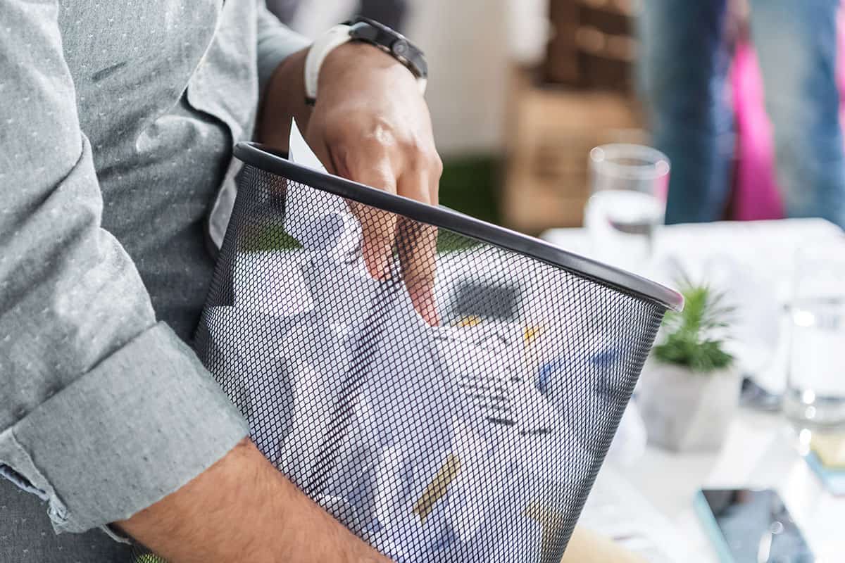 Trash Bin A person in a gray shirt is seen reaching into a black mesh trash can filled with crumpled papers.