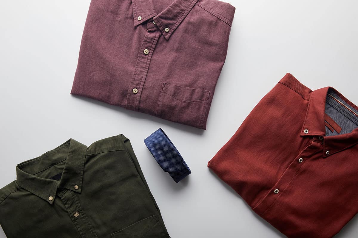 Assortment of Shirts Three neatly folded shirts in different colors: a maroon shirt, a burnt orange shirt, and an olive green shirt, accompanied by a navy blue tie, all arranged on a light gray background.