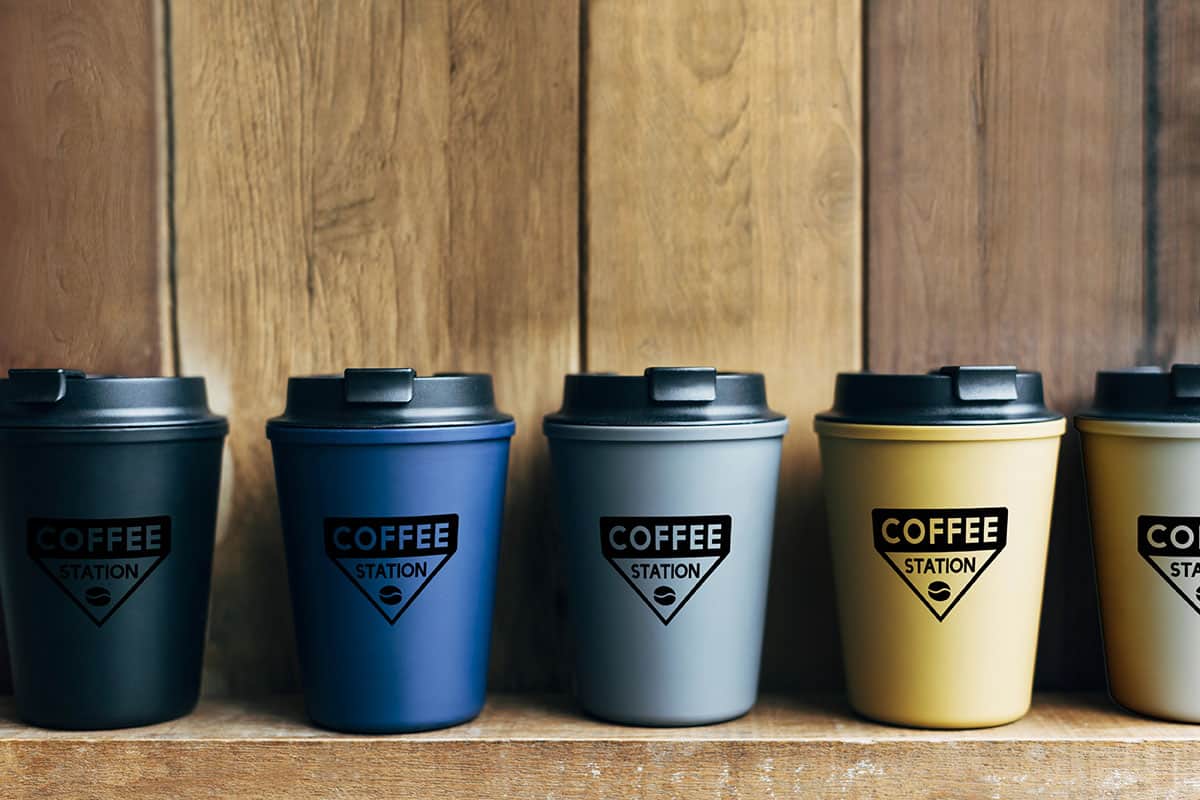 A row of four coffee cups in varying colors—black, blue, gray, and yellow—each with a black logo that reads "COFFEE STATION," set against a wooden background.