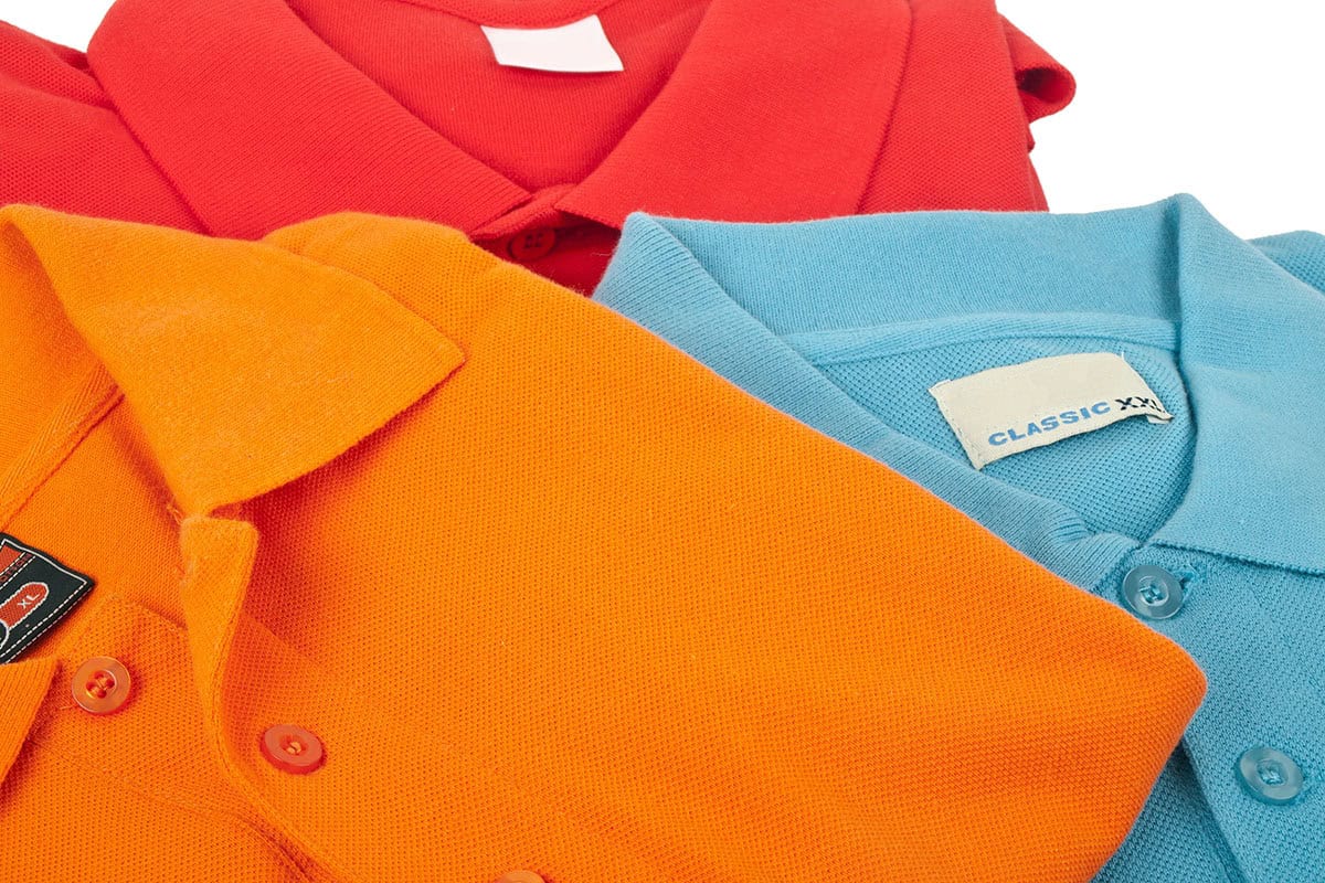 A close-up image of three polo shirts in vibrant colors: red, orange, and light blue, showcasing their collars and buttons. The labels indicate sizes and styles.