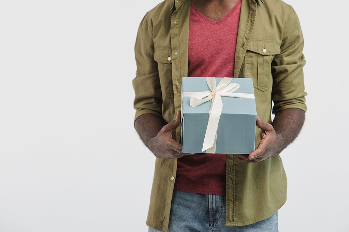 Gift Ideas A person holding a neatly wrapped blue gift box with a cream-colored ribbon, wearing a green shirt over a red t-shirt and blue jeans.
