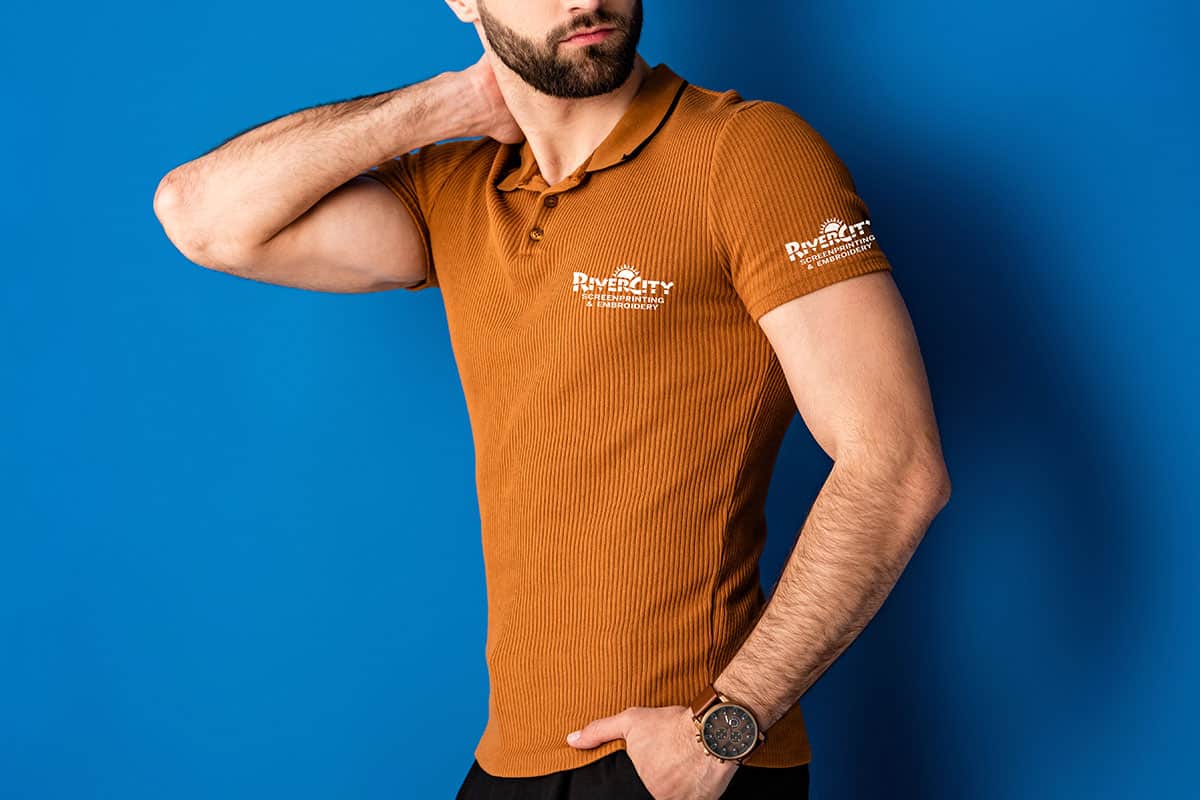 A man in a brown ribbed polo shirt stands against a blue background, with one hand on his neck and the other resting at his side. He is wearing a watch on his left wrist, and the shirt features white text on the left chest and right sleeve.