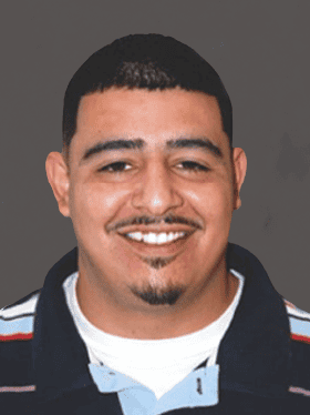 A smiling man with short dark hair, wearing a black and blue striped shirt, stands against a gray background.