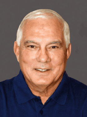 A smiling middle-aged man with white hair, wearing a navy blue polo shirt, against a grey background.
