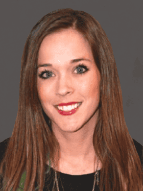 A smiling woman with long brown hair and bright blue eyes, wearing a black top and a necklace, against a gray background.