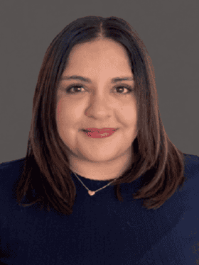 A woman with long, straight, dark hair and a warm smile, wearing a navy blue top and a delicate necklace, poses against a neutral background.