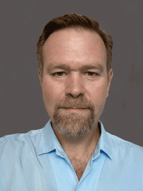 A man with a light brown beard and mustache, wearing a light blue shirt, poses against a neutral gray background.