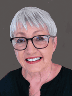 A smiling older woman with short, silver hair and glasses, wearing a black top, against a grey background.