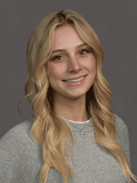 A young woman with long, wavy blonde hair smiles warmly at the camera, wearing a light gray sweater. The background is a soft gray.