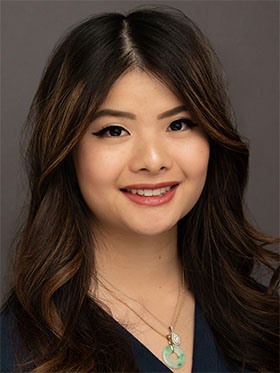 A smiling woman with long, wavy dark hair, wearing a blue top and a necklace, against a neutral background.