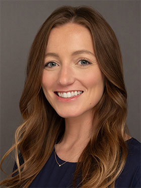 A smiling woman with long, wavy brown hair and blue eyes, wearing a dark top, against a gray background.