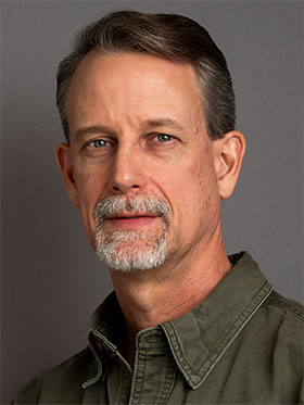 A man with short gray hair and a goatee, wearing a collared olive green shirt, looking directly at the camera with a neutral expression against a gray background.