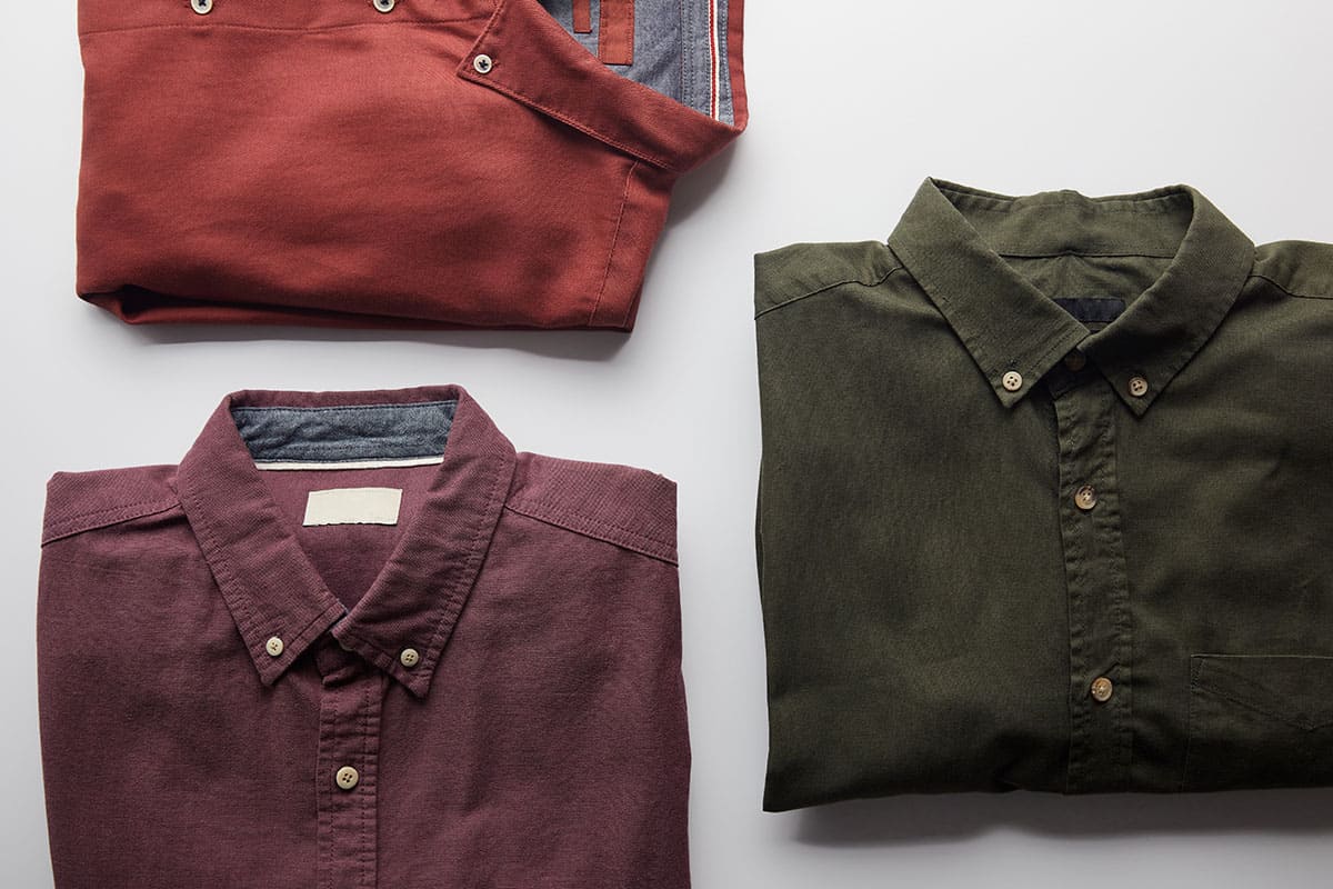 Three neatly folded shirts displayed on a light surface. The top shirt is a deep red color, the center shirt is a burgundy with a light blue collar, and the bottom shirt is olive green, all featuring buttoned collars.