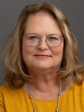 A middle-aged woman with wavy brown hair wearing glasses and a yellow shirt, smiling warmly at the camera.