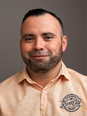 A smiling man with short, dark hair and a beard, wearing a light peach collared shirt with a logo on the left side, against a neutral background.