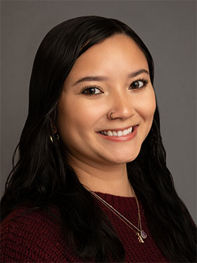 A smiling woman with long, dark hair, wearing a maroon sweater and multiple necklaces, poses against a gray background.