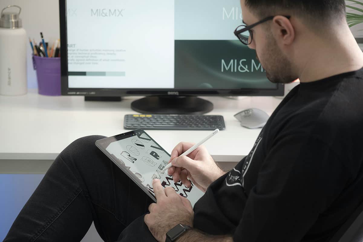 A person sitting at a desk uses a digital tablet with a stylus to create designs, while a computer monitor displays text related to "MI&MX." There are art supplies in a container and a water bottle visible on the desk.