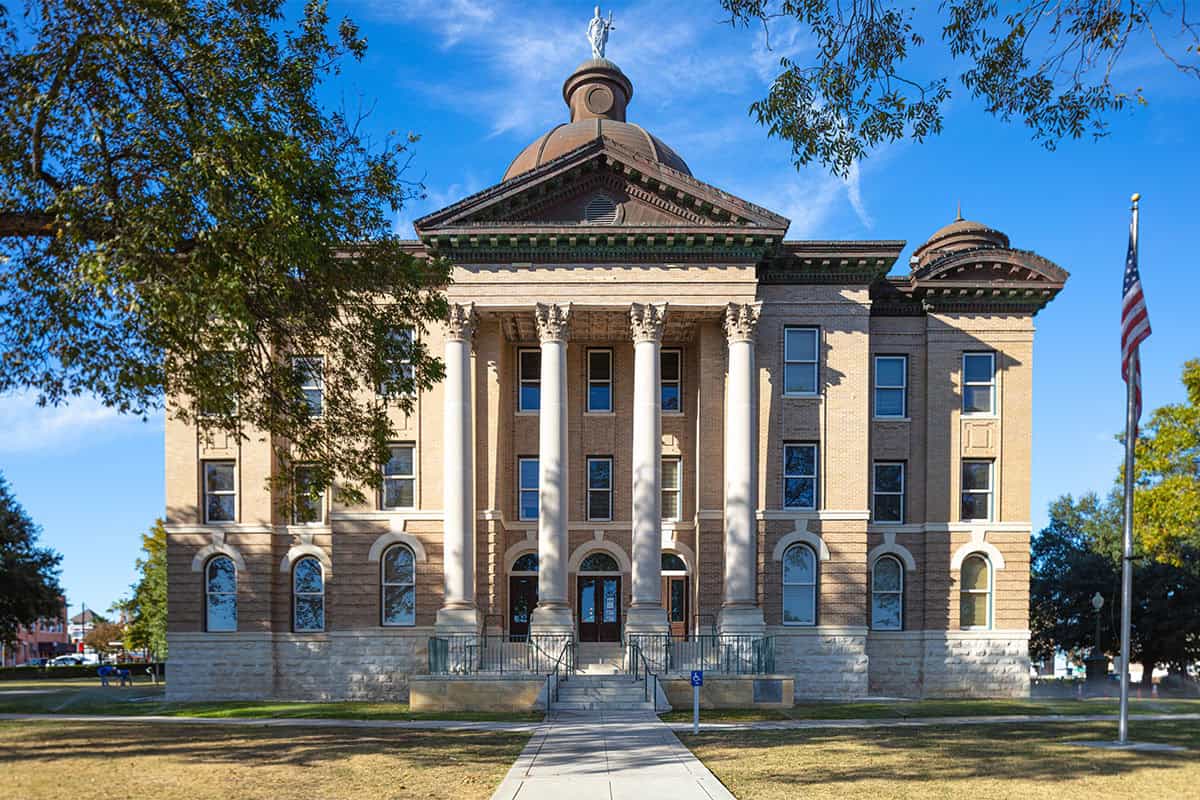 Historic courthouse building with a domed roof, featuring large columns and detailed architectural elements, surrounded by greenery and a clear blue sky.