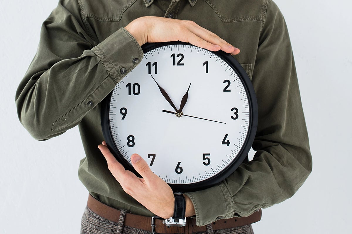 Holding Clock A person in a green shirt holds a large wall clock close to their body, showing the time at approximately 11:10. The background is plain and light-colored.