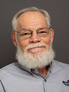 A smiling older man with a white beard and glasses, wearing a light blue shirt, poses against a gray background.