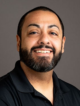 A smiling man with a beard, wearing a black polo shirt, poses against a neutral background.