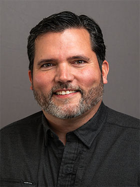 A smiling man with dark hair and a beard, wearing a black button-up shirt, poses against a gray background.