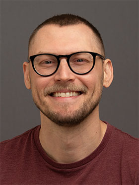Smiling man with short hair and glasses, wearing a maroon shirt, against a neutral gray background.
