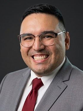 A smiling man wearing glasses, a gray suit jacket, and a red tie, against a dark background.