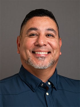 A smiling man with short dark hair and a goatee, wearing a dark blue collared shirt, against a grey background.