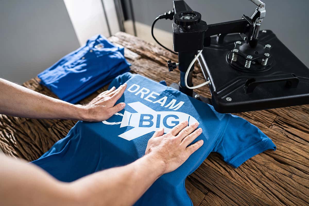 A person presses a blue t-shirt with the text "DREAM BIG" on a wooden table, using a heat press machine. Several folded blue shirts are visible in the background.