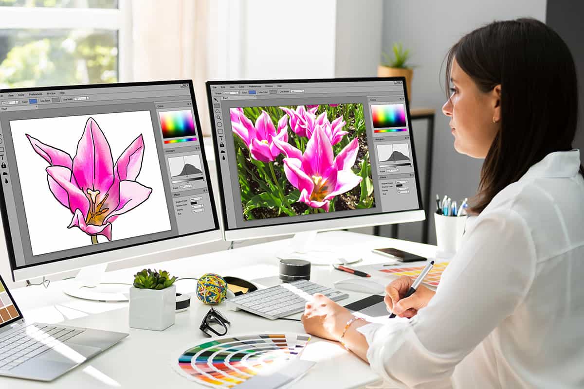 A woman sitting at a desk, focused on digital artwork, with two computer monitors displaying a pink flower illustration and a photograph of pink flowers. Color palettes and design tools are scattered around her workspace.